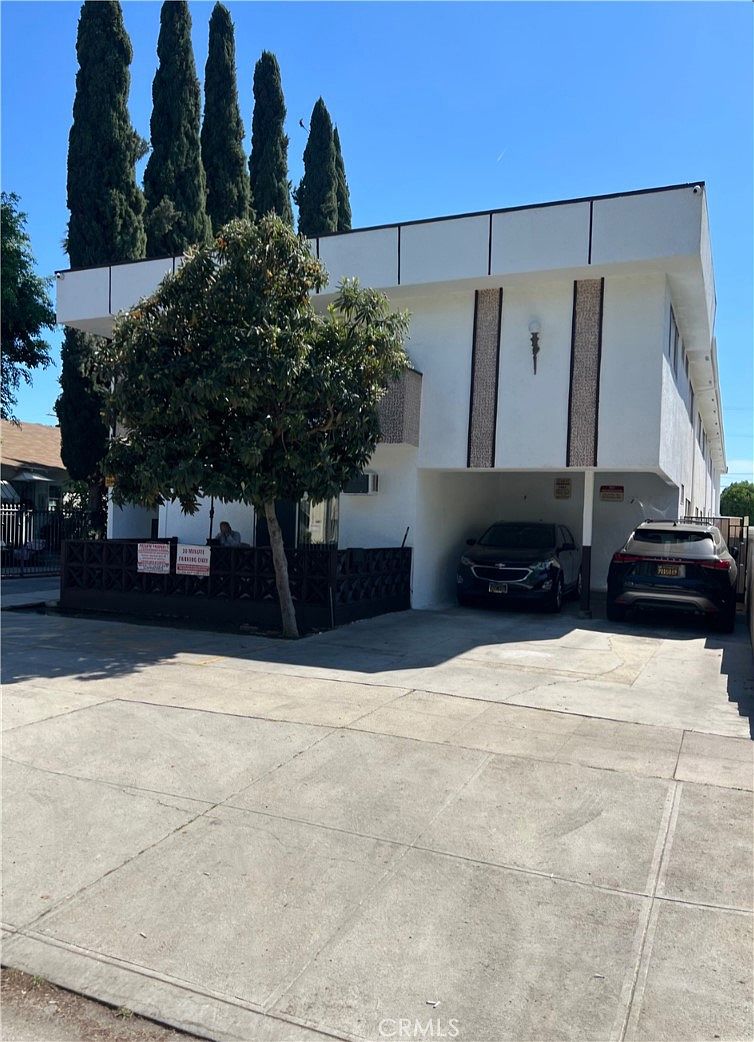 Three-quarter view of 14218 Sylvan St, a 1964 stucco apartment building in Sherman Oaks framed by mature Italian cypress trees, with mid-century stone-textured exterior panels and bracketed eaves